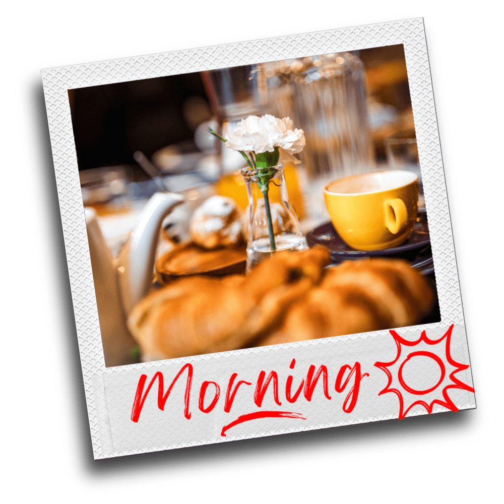 Polaroid photo of a breakfast table with a white flower in a vase, a yellow coffee cup, and croissants, captioned "Morning" with a sun doodle in red lipstick script lettering.